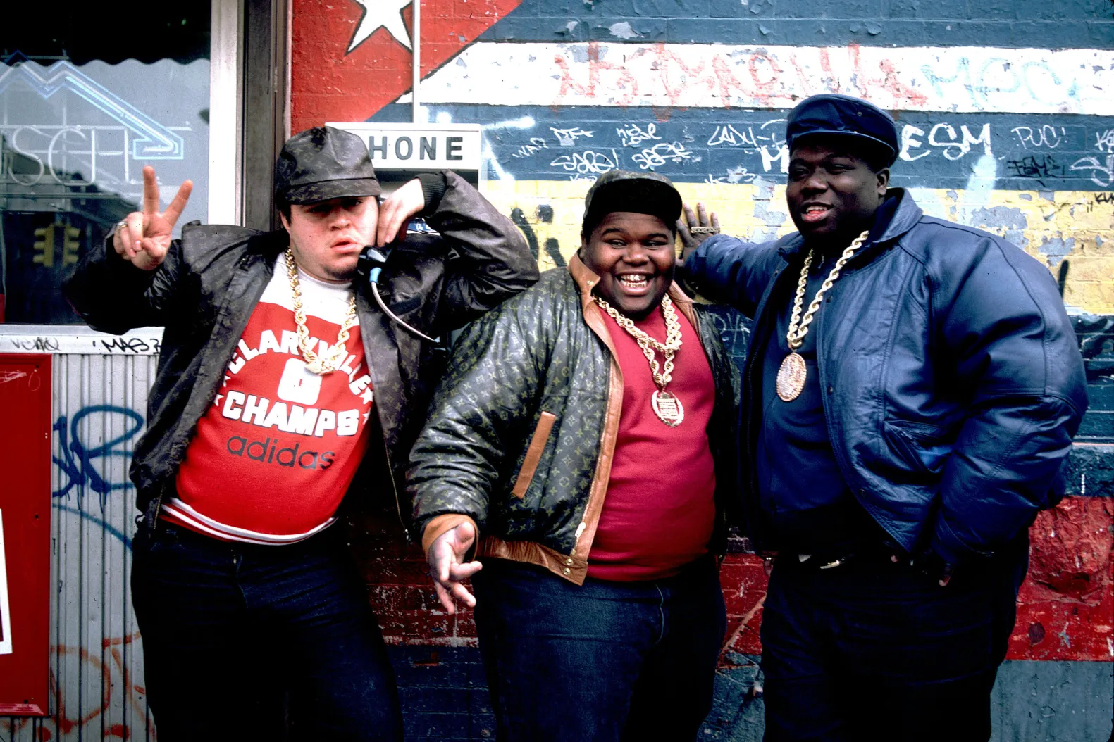 the fat boys posing in front of a colorful wall wearing jeans, hats, leather jackets, and large gold chains