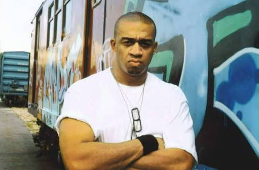 Artist, Lord Have Mercy, standing in front of a graffiti-painted subway car in a white t-shirt, arms crossed
