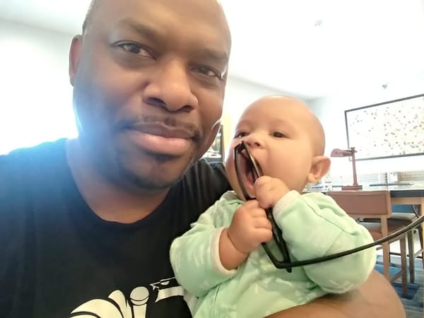 Selfie of me holding my infant daughter in a hotel lobby. I'm wearing a black t-shirt with a while logo. She is wearing light green onesie while attempting to put my glasses into her mouth