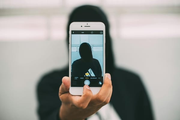 Hooded figure in black holding a cellphone in front of them with their own image displayed in the phone's camera view