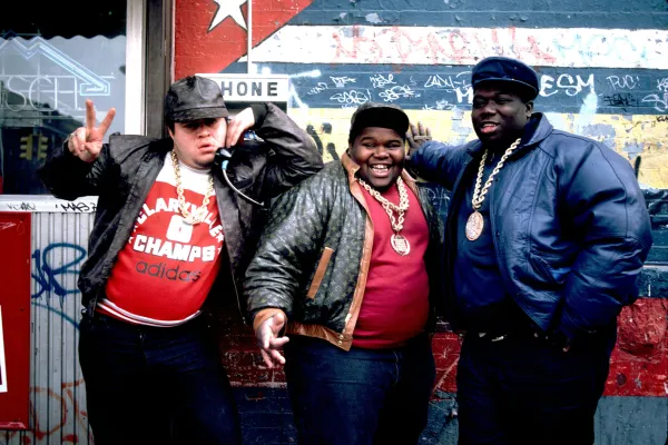 the fat boys posing in front of a colorful wall wearing jeans, hats, leather jackets, and large gold chains