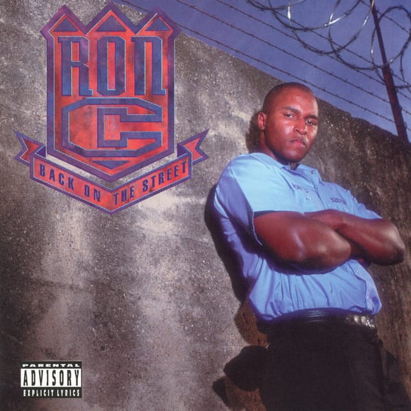 Album cover for Ron C "Back on the Street." Ron C is wearing black pants and a blue prison-style shirt with arms folder, leaning against a concrete wall with barbed wire